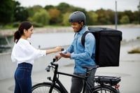 This photo shows an African-American courier standing next to his black e-bike. He is wearing a blue, long-sleeve, denim, button-front shirt; charcoal gray chino pants; a black bike helmet; and a large, black, box-shaped backpack. His right hand rests on the right handlebar. His left hand holds a white contactless payment device. A smiling Hispanic woman stands in front of him holding her credit card over the payment terminal. Dressed in a loose white blouse and dark slender jeans, she appears to be paying for a delivery. E-bikes like this courier’s transportation typically weigh more than a conventional bike, due to the motor and lithium-ion battery. Courier companies need to consider risks associated with lithium-ion batteries when selecting insurance.