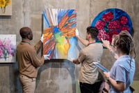 This photo features two men and one woman involved in hanging a painting at a museum. The men each hold one side of the painting against a grey display wall. Both are wearing white cotton archival gloves to protect against getting fingerprints on the artwork. It’s an abstract acrylic painting featuring various shapes in orange, blue, green, and white. The balding man on the left is African-American with tightly cropped hair. He’s wearing khaki pants, a camel-colored long-sleeve turtleneck shirt, and gold wireframe glasses. The man on the right is Caucasian with short, dark brown hair. He’s wearing a tan short-sleeve t-shirt and black slacks. In the foreground of the photo stands a white woman facing away from the viewer. She has ash-blond dreadlocks and rectangular brown glasses. Her left hand holds an iPad. Her right hand points at the painting, and she appears to be providing instructions to the two men. She is a fine art courier who helps protect the condition and value of artwork by ensuring no one takes shortcuts during packing, transportation, or installation. 
