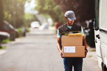 This photo shows a last-mile delivery driver in a brown vest and gray baseball cap. He is walking and carrying a stack of two brown boxes with some envelopes on top. His white delivery van sits with the back door open at the side of a residential street. Auto liability premiums for last-mile delivery companies have increased dramatically over the past twelve years. As a result, these businesses are exploring whether captives can help with insurance cost control.
