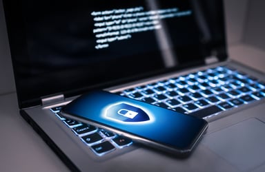 This photo shows a gray laptop with a black screen. Faint blue light emanates from the backlit keyboard. On the screen is a hacker’s malicious code. A slim gray cell phone rests on the keyboard. The cell phone’s screen features a dark blue gradient background with a shield shape above the midline. In the shield is a white padlock. A thick band of white light surrounds the shield. This decorative cybersecurity image reminds small businesses to consider the threat of cyberattacks and the need for cyber insurance.