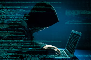 This image shows a side view of a computer hacker in a dark hoodie hunched over the keyboard of a laptop. The room is dark — navy blue with only the faintest hint of light. You cannot see the hacker’s face, adding to the sinister feel of the photo. Superimposed on the image is a blur of computer code and data. You get the sense this hacker is in the middle of a crime. Cybercrime against individuals and families has become increasingly prevalent. So, people are starting to ask, “Do I need personal cyber insurance?”