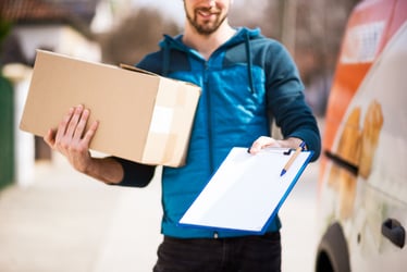 A  last-mile delivery man with a box in one arm and clipboard in the other making a delivery to a house.