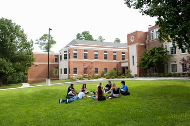 This photo shows eight college students meeting in a circle on a green lawn in front of an academic building. Getting property insurance for college and university buildings like this one has become more expensive, particularly in locations prone to catastrophic weather events. Investing in risk mitigation can help higher ed institutions reduce the impact of the hard property insurance market. 