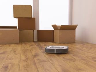 This photo shows a nearly empty room white room with two opaque floor-to-ceiling windows in the background. Six moving boxes sit atop a brown faux-wood floor. Three of the six are closed and in a stack. The remaining three are directly on the floor. The flaps remain open on one, as if the homeowner is still packing. In the foreground is a robotic vacuum cleaner. The lithium batteries that power household tools like this vacuum can pose a hazard during relocation. For this reason, it’s vital to follow the mover’s guidelines on lithium-ion battery safety, even if this means recycling your current batteries and purchasing new ones at your destination.