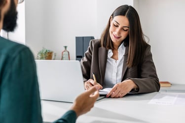 This image depicts a professional woman engaged in a meeting with a man, both sitting at a table. The woman is actively writing in her notebook, symbolizing discussions on the impact of pay range disclosure statutes, potential risks of lawsuits, and considerations for insurance coverage.