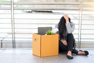 This photo shows a downcast businesswoman sitting on the floor with a box of belongings. She appears to have lost her job. Layoffs lead to a range of emotions, which can include sadness and anger. If handled poorly, layoffs can result in claims of discrimination, lawsuits, and employment practices liability insurance (EPLI) issues.  