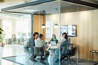 This photo shows six businesspeople sitting around a small rectangular conference table — three men and three women. The conference room has floor-to-ceiling windows on three sides, giving it a “fish tank” feel. Vertical blond wood panels cover the fourth wall. Seated in modern black chairs with low backs and black metal legs, the meeting participants are leaning in, listening to a woman in a gray pants suit and short gray bob haircut at the righthand end of the table. Decisions made in meetings like this sometimes lead to lawsuits. Startup companies can be particularly vulnerable to legal action due to potential lack of knowledge about employment law. D&O insurance can help protect startups from unexpected legal expenses. 