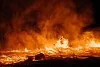 This photo shows a wildfire, sparks, and smoke at night. Trees are in the background, and one is fully engulfed in flames. Smoldering wood sits in the foreground, suggesting the fire is moving away from the viewer to consume the fuels in the background. Fires like this are terrifying for nearby homeowners. Before disaster strikes, it’s important to create a detailed wildfire evacuation plan for your family’s safety. 