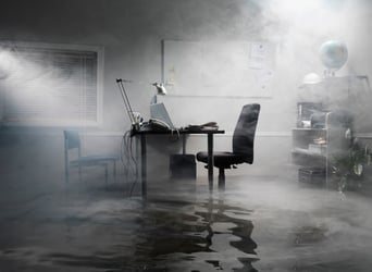 This mostly black and white photo shows an office with several inches of water covering the floor. Wispy smoke hangs in the air, suggesting a fire not just a flood. In the background is a whiteboard and a window with mini blinds. On a black desk sits a laptop, monitor, telephone, and desk lamp, with power cords in the water. Under the desk, a computer tower is partially submerged. On the right, a black desk chair faces the equipment. Behind the desk chair, on the right wall, is a potted plant and a shelving unit with miscellaneous office equipment and supplies. On the other side of the desk is a simple guest chair with a black metal frame and teal upholstery. When a catastrophic event renders an office unusable, business interruption insurance can help protect the balance sheet. However, it’s important to check what your BI policy does and doesn’t cover — to make sure you have the protection you need. 