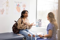  A young African-American woman student engages in a conversation with a medical professional in a health clinic.  Educational posters in the background suggest the setting is a student health center, where  medical care and student health insurance topics are commonly addressed.