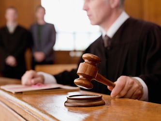 This image shows a judge in a black robe sitting at the bench, with a wood gavel in his left hand raised as if he’s about to pronounce judgement. His right-hand rests on two file folders. In the background, two jurors stand in the jury box. No faces show, and the people are blurry, so the focus is on the gavel. Last-mile delivery companies find themselves in courtrooms like this one, defending against negligence claims. Opportunistic investors are funding these lawsuits, angling for a share of the settlement. This so-called “third-party litigation funding” is driving up insurance rates for the entire transportation industry. 