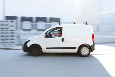 This photo shows a tiny white delivery van driving in an industrial area. The van is clean and shiny with no business name on it. Blurred in the background is a building with four loading docks. Businesses that deliver cannabis products use unmarked vehicles like this one to reduce the risk of theft. Because of cannabis banking restrictions, drivers often carry more cash than in other industries, making them a robbery target. This is one of several reasons insurance companies have been slow to develop insurance products to protect cannabis transportation. 