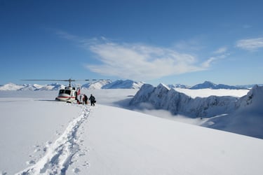 This photo shows a helicopter on a remote snowy mountaintop. Two people are preparing to board, with ski gear planted nearby in the snow. The helicopter has a white fuselage with a decorative red stripe from nose to tail, and a red tail. Above the deep snow is a blue sky. Adventurers like these need to plan their travel insurance carefully. If the weather or an accident derails travel plans, do you have adequate coverage for the resulting expenses?