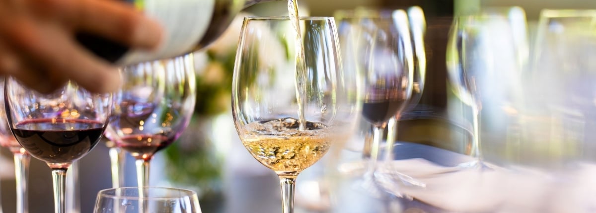 Hand pouring white wine into a glass, surrounded by other glasses of red and white wine, set against a blurred backdrop of greenery and soft lighting, hinting at an elegant outdoor event or vineyard tasting. Insurance for wineries is essential risk protection for wineries who serve alcohol.