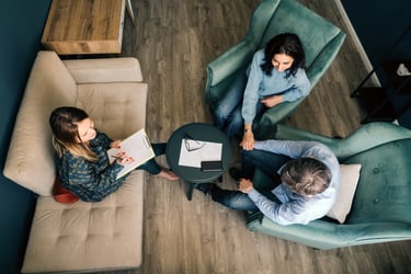 In this photo, there is an overhead view of three individuals in what appears to be a professional setting. There is a small round table in the center with a pair of glasses, some papers, and a smartphone on it. The setting suggests a relaxed yet professional meeting or consultation, possibly in a creative or counseling context.