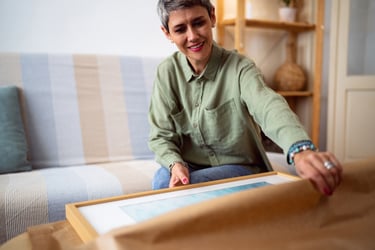 A middle-aged female with short gray hair, who is seated, unwrapping brown paper from a framed artwork. Fine art insurance for artwork in transit is vital, because unfortunately, transportation damage is common.