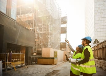 This photo shows a side view of two engineering professionals standing at a construction site. Both are wearing yellow safety vests with reflective stripes around the lower rib cage and hips. Both also are wearing blue construction hard hats. The person on the right is pointing up at the construction project while holding a tablet in their left hand. The person on the left is holding a roll of paper that may be architectural drawings. The background includes construction fencing, scaffolding, and in-progress buildings. In any construction project, such as the one shown in this image, an A&E firm can face setbacks — such as workplace injuries or supply chain delays. It’s important to alert your professional liability insurance carrier early about any circumstances with the potential to become a claim. 