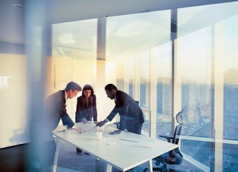 A team of collogues gather around a table in an office located within a high rise office building discussing pharmacy benefit plan designs.
