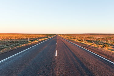A straight road stretching towards the horizon in the distance. The road is asphalt and appears to have two lanes in each direction, with a wide dividing line in the center. There are no cars or other vehicles on the road.