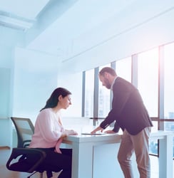 A white female human resources professional sitting at a desk is meeting with a white financial professional in an office setting with sun shinning through the windows.