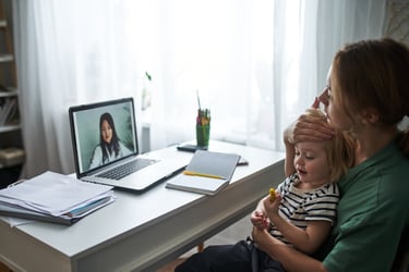 A mother holds her sick child during a telehealth appointment with a doctor. The doctor is visible on a laptop computer on the desk they are sitting at.