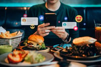 A person dining in a restaurant and engaging with their smartphone. They are surrounded by food - a burger, fries, and what looks like a grilled shrimp dish. Overlaid on the image are various social media icons and emojis, like hearts, thumbs-up, and smiley faces, suggesting that the person might be interacting with a social media platform.