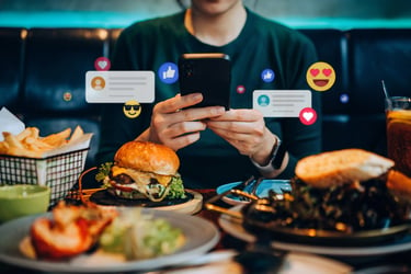 A person dining in a restaurant and engaging with their smartphone. They are surrounded by food - a burger, fries, and what looks like a grilled shrimp dish. Overlaid on the image are various social media icons and emojis, like hearts, thumbs-up, and smiley faces, suggesting that the person might be interacting with a social media platform.