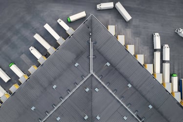 A large warehouse with a vast parking lot filled with semi-trucks. The trucks are parked in neat rows, with some backed up to loading docks. The warehouse has a large flat roof and multiple garage doors. Relevant to this photo: A freight broker is a company that acts as an intermediary between shippers and carriers. They arrange for the transportation of goods, but they do not own or operate their own trucks.