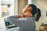 A female remote worker experiencing neck fatigue, displayed by her grabbing the back of her neck with her left arm.  Worker's comp insurers have seen an increase in upper extremity (hands and wrists) and back injuries, as well as neck and shoulder issue.